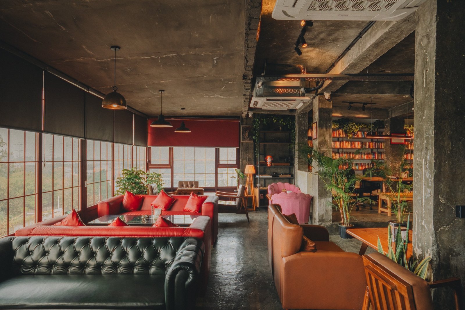 Wide view of the café lounge: tufted sofas, red cushions, bookshelves and warm pendant lights